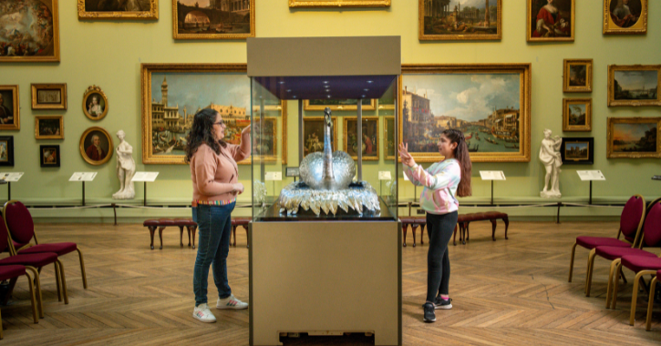 Mother and daughter standing either side of Silver Swan Automaton at The Bowes Museum with painting covering the walls in the background. 